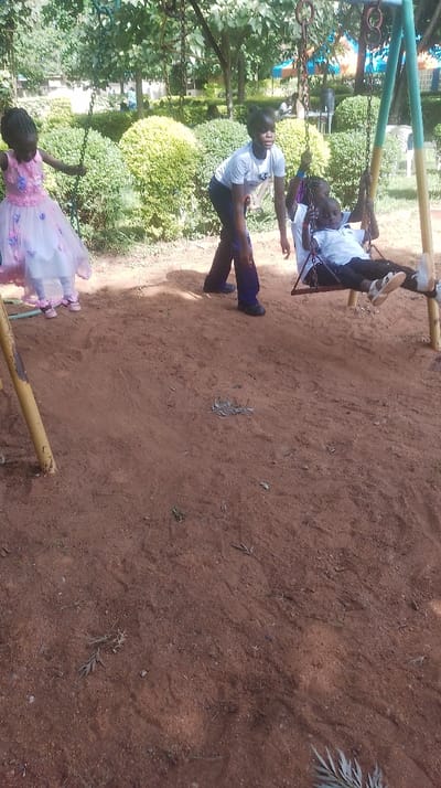 The scene depicts an outdoor playground in Siaya Township ward, Kenya, during daylight hours. Three individuals are present on reddish-brown dirt ground. On the left, a young female child, estimated 4-6 years old, wears a pink and white tiered dress with floral embellishments and pink sandals. She stands near a yellow swing post, holding one of the swing chains. In the center, an older female, estimated 12-16 years old, wears a light grey t-shirt with a graphic print and dark trousers with dark shoes. She is bending forward, actively pushing a swing. The swing, constructed with a red-brown seat and chains, contains a second young child, estimated 3-5 years old, who is reclining. This child wears a white collared shirt, dark trousers, and light-colored sandals. The swing set posts are yellow and teal/green. The background features manicured green bushes, providing a hedge-like barrier, and numerous trees with green foliage. Faint architectural elements and white chairs are partially visible behind the bushes.