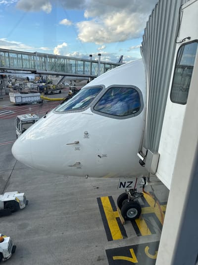 The image shows the nose of an Air New Zealand (NZ) aircraft parked at a gate, likely in Le Mesnil-Amelot, France, given the Air France gate visible in the background. The aircraft is connected to a jet bridge. Ground equipment, including tugs, are visible near the nose gear. The markings on the ground indicate the aircraft's position at gate 7. The sky with clouds is visible through the jet bridge windows.