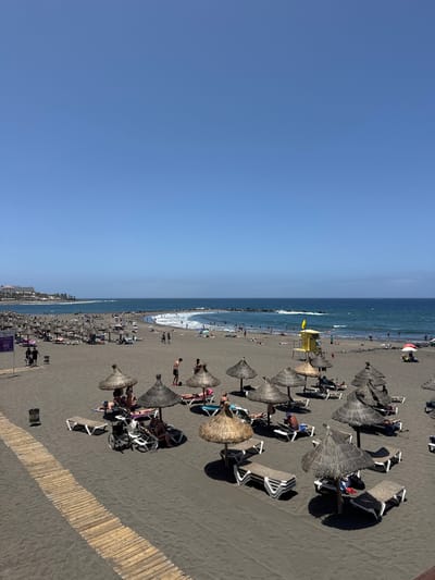 This is a wide shot of a black sand beach in Adeje, Spain, on a clear, sunny day. The sky is a vibrant, cloudless blue. The beach is populated with numerous sunbathers relaxing under straw umbrellas. Many lounge chairs are set up on the sand, some occupied by people, others empty. In the foreground, a wooden walkway runs diagonally across the sand. To the left, a metal trash can stands near the walkway. Further into the scene, people are swimming and wading in the blue ocean. Waves are breaking near a rocky breakwater that extends into the sea. A yellow lifeguard tower with a yellow flag is visible on the sand. The coastline stretches into the distance, with buildings and palm trees lining the shore.