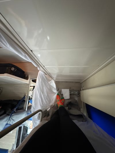 The scene is an interior view of a train sleeping carriage, consistent with *platskart* class coaches utilized in Russia, and is noted as potentially occurring in the vicinity of Gryazi, Russia. The perspective is from a lower bunk, looking towards the foot of the bed and the opposite side of the carriage.
In the immediate foreground, the legs and feet of an individual are visible, clad in black leggings and distinctive orange socks adorned with green and red patterns, with the feet crossed at the ankles. The individual rests on a bunk covered by white sheets and a grey blanket. To the immediate left, a white privacy curtain is partially drawn, suspended from a ceiling-mounted track. To the right, a white wall panel features a wire storage basket containing a folded white towel, positioned above a blue panel section.
The carriage ceiling is white and glossy. In the background on the left, a black wheeled suitcase with visible wheels is stowed on an upper bunk, and a lower bunk is partially visible with dark items. Metal safety rails are integrated into the bunk structures. The interior walls beyond the immediate bunk area are light brown.