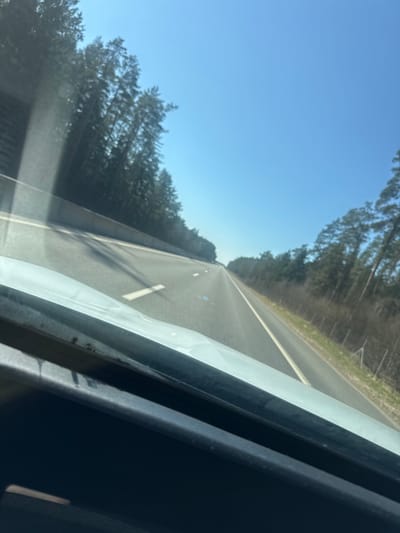 The image, taken from inside a moving vehicle, shows a multi-lane highway cutting through a forest. The road is mostly straight, with white lane markings and intermittent white dashes. Trees line both sides of the highway. The sky is blue and clear. The interior of the car is partially visible on the right side of the frame. Based on the landscape, this image could be located in the wooded areas of Іўеўскі сельскі Савет, Belarus.
