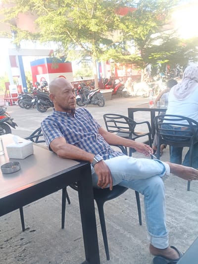 The image depicts an outdoor scene at a gas station, likely in Geulumpangsulu Timu, Indonesia, given the context provided. In the foreground, a bald, middle-aged man with weathered skin is seated at a dark table. He is wearing a blue and white checkered shirt and light blue jeans, with one leg crossed over the other. A silver watch is visible on his left wrist. On the table in front of him are a tissue box and an ashtray with cigarette butts. To his right, two black chairs are partially visible. In the background, several motorcycles are parked under a covered area. Behind the motorcycles, a gas station structure with red and blue accents is prominent. Trees with green foliage are visible, and sunlight illuminates the scene, casting shadows. A person wearing a white headscarf and a light-colored shirt is seated at another table in the mid-ground. The ground surface is concrete with some visible wear and tear. The overall impression is of a casual setting at a gas station during daylight hours.