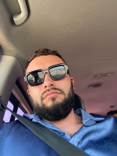 The image shows a close-up, selfie-style shot of a man with dark hair, a beard, and sunglasses, sitting in the passenger seat of a vehicle. The man is wearing a blue button-down shirt with a repeating pattern. The interior of the car is visible, including the ceiling with a grab handle and lights. The man's sunglasses reflect a road with passing cars. The provided location of Koshkeldy, Russia, is not directly discernible from the visual information in the image.