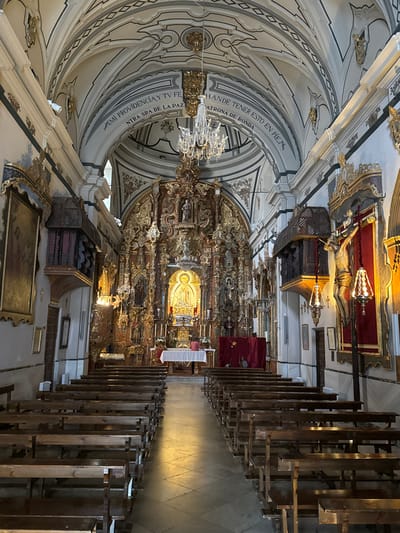 The image displays the interior of the Sanctuary of Nuestra Señora de la Paz, located in Ronda, Spain. The perspective is from the nave, looking directly towards the main altar.

The environment is a large hall with a vaulted ceiling painted in white with grey and black geometric and curvilinear patterns. Several crystal chandeliers are suspended from the ceiling. The walls are white and extensively adorned with gold-gilded ornamentation and cornices. The floor is covered in light-colored, patterned tiles, with a central aisle leading to the altar. Dark wooden pews are arranged in parallel rows on both sides of this aisle; most appear unoccupied.

At the far end, a highly ornate, gilded Baroque altarpiece (reredos) dominates the scene. It features multiple tiers and niches, each containing statues. A central figure within a niche at the lower-middle section of the altarpiece is illuminated with a warm golden glow. In front of this altarpiece, a table draped in white cloth serves as the altar, decorated with white floral arrangements and candles.

Two individuals are present in the sanctuary area to the right of the altar. One person, wearing a reddish top, stands positioned behind the altar table, facing towards the right side of the image. Another individual, dressed in darker clothing, is seated on a low structure further back on the right, partially obscured by a draped red fabric. Behind this red fabric, indistinct forms are visible.

To the right of the altar, a large crucifix depicting a wooden corpus of Christ is mounted against a red fabric panel, flanked by decorative hanging lamps. Above the pews on the right wall, a dark carved wooden structure is affixed. On the left wall, a large painting with a prominent gold frame is visible. A small, white security camera is mounted high on the left wall, directed towards the altar.