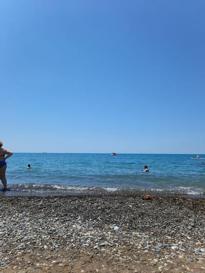 The image captures a serene beach scene in Krinitsa, Russia, under a clear blue sky. The foreground is dominated by a pebble beach, with the gentle waves of the Black Sea lapping at the shore. A few people are visible in the water, enjoying the summer day. In the middle ground, a small boat with people on board can be seen, and further out, a person is paddleboarding. To the left, a woman stands on the beach, facing the sea. The overall impression is one of a peaceful and pleasant day at the seaside.