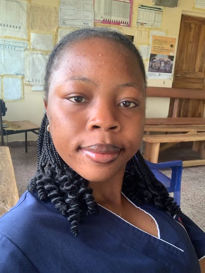 This is a vertical, close-up selfie of a young Black woman. She is wearing a dark blue uniform with white trim. Her dark hair is braided and styled in twists. She is looking at the camera with a slight smile. Her skin appears to have some minor blemishes. The background is out of focus, but it appears to be an indoor setting, possibly an office or clinic, with walls covered in charts and papers. There is also some wooden furniture visible.

One of the papers on the wall has text, but it is too blurry to read. The scene appears to be well-lit, suggesting it is daytime. The woman's expression is relaxed and friendly.

Based on the provided location context, this image was taken in Reak, Nigeria.