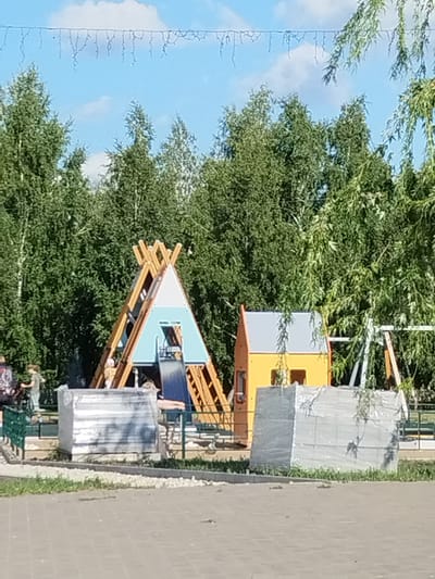 An outdoor children's playground is depicted in an urban park setting, likely in Рощинский, Russia. In the foreground, a paved walkway transitions to a gravel and grass area. Two large, rectangular objects wrapped in grey plastic sheeting are positioned on the grass. Behind these objects, a colorful playground structure is visible, featuring a teepee-like climbing frame with a blue section and a yellow playhouse. To the left of the teepee, a child is seen on a bicycle with their guardian standing nearby. Further to the right, a swing set frame is partially visible. The background is dominated by dense green trees, and a clear blue sky with a few scattered clouds is overhead. A string of decorative lights is suspended across the top of the image.