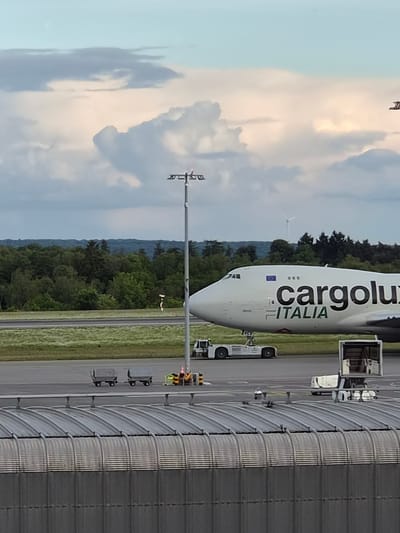 The image shows a Cargolux Italia cargo plane at Luxembourg Airport (LUX) in Niederanven, Luxembourg. The plane is parked on the tarmac next to a metallic building. A communication tower is visible in the foreground. Beyond the tarmac is a backdrop of trees and distant hills. The sky is partly cloudy.