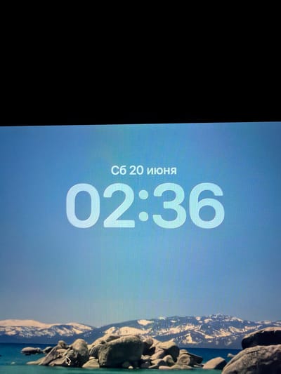 The image displays a digital clock showing the time as 02:36. Above the time, the date is indicated as "сб 20 июня" which translates to "Saturday, June 20". The clock is superimposed on a serene landscape featuring a body of water, numerous large rocks in the foreground, and a snow-capped mountain range in the background under a clear blue sky. There is a mouse cursor visible on one of the rocks in the lower left portion of the image. The image does not depict any specific location in the city of Razumnoye, Russia, or any people.