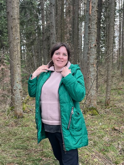 A woman is standing in a forest in Bol'shakino, Russia. She is wearing a green puffer jacket over a light pink hoodie and black pants. The forest is characterized by tall, thin trees and ground cover of dry grass and twigs.