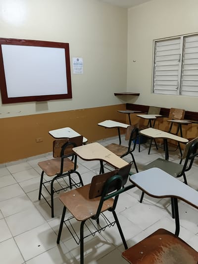 This image depicts an empty classroom in Puerto Plata, Dominican Republic. The room contains rows of desks with attached chairs, most of which are made of wood and metal. A large, blank whiteboard with a brown frame is mounted on the front wall. To the right of the whiteboard, a small sign with Spanish text is displayed. The walls are painted in two tones: cream on the upper portion and a mustard yellow on the lower portion. A louvered window with white blinds is visible on the right wall. The floor is tiled with white squares. The scene suggests a typical educational setting, currently unoccupied.