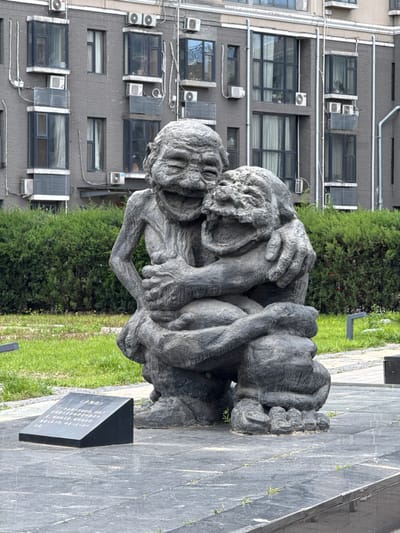Here is a description of the image:

This image shows a stone sculpture of two intertwined human figures, likely depicting the elderly, positioned in an outdoor setting. The sculpture is situated on a paved area adjacent to a patch of grass and a dense green hedge. Behind the hedge, a multi-story building with numerous windows and air conditioning units is visible. To the right of the sculpture, a dark, upright stone plaque with text in Chinese characters is present.

This photograph was taken in the city of Changping District, China.