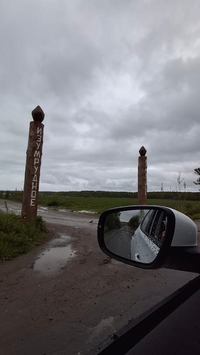 A slightly high-angle, close-up outdoor shot depicts a gray car's side mirror capturing a view of a dirt road flanked by green vegetation and a forest edge under a cloudy sky. A weathered wooden signpost with a pointed top, inscribed with "NEMPAVAHOE" in white block letters, is visible both directly and in the mirror's reflection, indicating a location in Nempa, Russia. The road surface is wet, with puddles reflecting the overcast sky. The car's side panel is partially visible in the right edge of the frame.