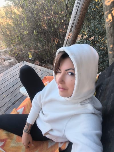 An outdoor, high-angle image, taken in Shaalvim, Israel, depicts a female subject seated on a weathered wooden platform. The individual, with short brown hair, wears a light-colored hooded sweatshirt with the hood worn up, black leggings, and white sneakers on her left foot. Her right eye is open and facing the camera, adorned with blue eyeliner and small glitter applications. Her left eye is winked shut, and her tongue is slightly extended. A nose piercing is visible on her left nostril, and a tattoo is present on her right forearm. She is seated on an orange and white geometric-patterned blanket covering the wooden platform. The background consists of dense, green, and sparse foliage with exposed rocky terrain. Two weathered wooden posts are visible on the right side, with metal chains hanging near the left post. The scene is illuminated by warm, directional sunlight.