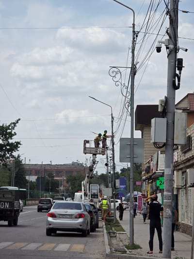 The image, taken in Talgar, Kazakhstan, depicts a street scene with overhead power lines and a utility pole. Two individuals wearing safety vests and helmets are visible in a lift basket attached to a crane, appearing to be working on the power lines. Several cars are parked or driving on the street, including a silver Toyota sedan with license plate "557 BG 11 05" and a dark colored truck in the foreground. Buildings line the street, suggesting an urban or suburban environment. The sky is overcast with clouds, indicating a cloudy day. A digital display showing the number "2" in green is visible on a pole. The overall impression is of ongoing utility work in a residential or commercial area.