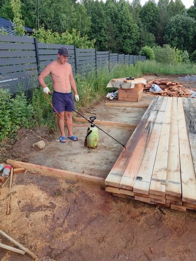 A shirtless young man wearing a dark baseball cap, white gloves, and blue flip-flops stands in a construction area, holding a spray bottle with a long wand. He is spraying a stack of wooden planks laid out on the ground. The planks are a light wood color, with some showing dark staining. The ground is a mixture of dirt, wood shavings, and some muddy patches.
Behind the man, a dark grey, slatted fence runs horizontally across the frame. Beyond the fence, a dense forest of green trees is visible, with some lighter green foliage in the foreground. To the right of the man, a wooden crate covered with a canvas-like material is visible, with rolls of material next to it. A pile of cut lumber is also present in the background.
The scene appears to be outdoors, likely in a rural or semi-rural setting, given the presence of the forest and the unpaved ground. The lighting suggests it is daytime, possibly late afternoon, with the sun casting soft light. The man's posture and focus on the task imply he is engaged in a construction or preparation activity, possibly treating the wood. No visible text is present in the image. The overall mood is one of work in progress.