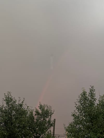 The image depicts a low-angle view of a hazy sky, with the tops of green trees framing the right and lower right portions of the frame. A faint, pale rainbow or atmospheric optical phenomenon is visible in the sky, horizontally across the middle of the image. The overall atmosphere is muted and diffused due to the haze. The presence of tree foliage and what appears to be a wooden pole with wires in the upper right corner suggests an outdoor, likely rural or suburban, setting. The time of day is ambiguous due to the overcast conditions, but the soft light could indicate dawn, dusk, or a heavily overcast day. There is no visible text in the image. The scene conveys a sense of quiet or perhaps a slightly melancholic atmosphere due to the muted colors and hazy conditions.