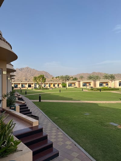 The image depicts a resort setting in Al Fiqait, United Arab Emirates. A row of multi-story buildings, possibly hotel rooms, flanks a well-manicured lawn. Behind the buildings, mountains are visible. The foreground features a set of stairs with a patterned walkway, and landscaping including plants. The sky is clear and blue. There are no people visible in the scene.