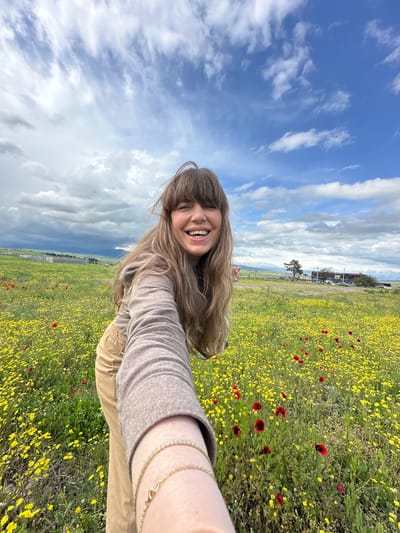 A woman is taking a selfie in a field of yellow and red flowers near Marneuli, Georgia. The sky is partly cloudy. A building, trees, and a fence are visible in the background. She is wearing a beige long-sleeved shirt and khaki pants. She is smiling. She has long brown hair with bangs. Two gold bracelets are visible on her left wrist.