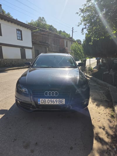 A black Audi sedan is parked on a paved street in Kaleytsa, Bulgaria. The license plate reads "OB 0969 BE". The car is oriented towards the viewer with the front end visible. Behind the car, traditional Bulgarian architecture is visible, including a white building with brown trim and stone accents. Power lines are visible overhead. Trees partially obscure the view. The sun is shining brightly. The street surface is asphalt.