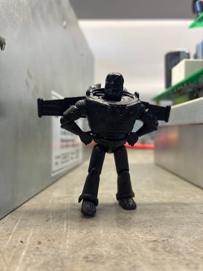 This media file features a close-up, slightly angled shot of a black, 3D-printed figurine, possibly a robot or action figure, lying on a textured, grey surface. The figurine is posed with its limbs bent and its body facing the camera. The surface it rests on appears to be a workbench or a similar utilitarian surface, showing signs of wear and tear, with scuffs and faint dark marks.
Behind the figurine, the background is out of focus. There is a glimpse of electronic components, including a green circuit board with white rectangular elements. The overall impression of the setting is that of a workshop or a space where electronic equipment is handled. The lighting suggests an indoor environment, likely artificial, with soft shadows. There is no discernible text visible in the image. The subject is the figurine itself, with the background providing context for its potential creation or use.