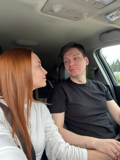 A man and woman are in a car. The man has short brown hair and is wearing a black t-shirt. The woman has long red hair and is wearing a white shirt. They are holding hands. The man has a tattoo on his arm. The location is Chastyye, Russia.
