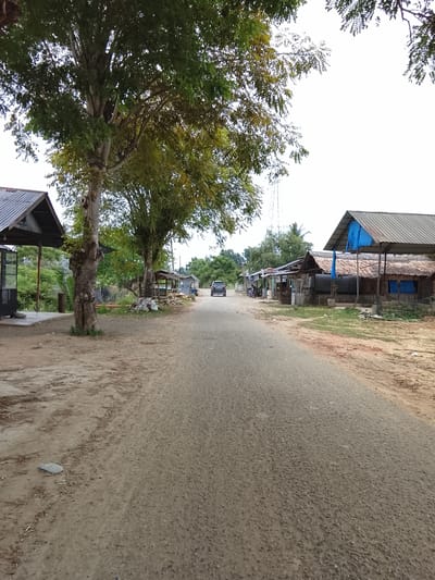 The image depicts a dusty, unpaved road in what appears to be a rural or semi-urban area, likely within Rawasari, Indonesia. Several structures line the right side of the road, some with corrugated metal roofs and others with thatched roofs, suggesting small businesses or residences. One structure on the far right has a blue tarp covering part of its roof. Trees with dense green foliage are prominent on the left side of the road, with one large, gnarled tree in the foreground. A small, dark-colored SUV is driving away from the viewer down the road. The environment is predominantly earthy tones, with scattered debris and vegetation visible along the roadside. The sky is overcast and bright white.