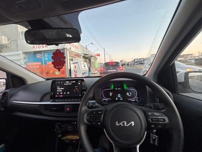 The image captures a first-person perspective from inside a stationary Kia vehicle, looking forward through the windshield. The time displayed on the car's infotainment screen is 8:00 AM on May 6th.
Inside the vehicle:
The black leather-wrapped steering wheel, bearing the silver KIA logo, dominates the lower center. Controls for media, phone, cruise control, and lane keeping assist are visible on the steering wheel spokes. A set of keys hangs from the steering column. The digital instrument cluster behind the steering wheel shows the vehicle is stationary (0 km/h), in neutral (N), with an odometer reading of 12,327 km. The headlights and lane keeping assist indicators are illuminated green. The infotainment screen displays "Bluetooth Audio" with the song "Thee Mou" by Mika Metaxas from the album "One of a Kind." Below the screen, the hazard light button is illuminated red. A red, leaf-shaped "air." brand air freshener, labeled "Energy," hangs from the rearview mirror.
Outside the vehicle:
The scene is an urban environment in the Ypsonas Municipality, Limassol District, Cyprus, specifically facing east on Leoforos Makariou III, near the intersection with Agiou Georgiou. Directly ahead, a dark gray Nissan vehicle with Cypriot license plate "KZK 760" is visible, its taillights illuminated. To the left (north side of the road), a white building complex houses a "KIOSK" with the partially visible name "ΕΥΡΟΠΗ ΔΕΡΒΙΣ." The kiosk's storefront features an advertisement for "PAPAFILIPOU ICE CREAM," displaying various flavors, along with "PSP" branding. Multiple commercial freezers branded "PARTY ICE" and "Party Ice cubes" are positioned outside the kiosk. Text such as "COFFEE" is also visible. Power lines traverse the clear, light blue sky above the road. On the right side of the road, and further down the street on the left, modern residential buildings line the street. No individuals are explicitly visible.