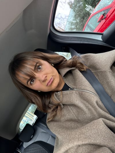 A female individual is depicted in a selfie taken inside a vehicle in Mougins, France. She has brown hair with bangs and brown eyes, wearing a light brown or beige textured jacket with a visible silver zipper over a dark garment. A dark grey seatbelt is diagonally positioned across her chest and shoulder. Her expression includes furrowed eyebrows and pursed lips. The vehicle interior shows dark upholstered seating behind her. Through the rear window, raindrops are visible on the glass, and a portion of a red car is discernible outside, accompanied by blurred green foliage and a paved surface, indicating an overcast and wet exterior environment.