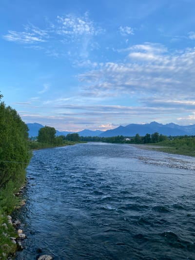 This media file depicts a scenic landscape view of a river in Novosnezhnaya, Russia, likely taken during the day. The image is oriented vertically.

The primary subjects are the natural elements of the scene. A wide, fast-flowing river with visible ripples and texture dominates the right side of the frame. Its banks are lined with green trees and large stones. In the background, a lush green forest borders the river, leading up to a range of majestic mountains, some of which appear to have snow-capped peaks. The sky is a clear blue, adorned with scattered, wispy white clouds that catch the light. The overall impression is one of natural beauty and tranquility.

There are no visible people or specific objects of interest beyond the natural landscape itself. No activity or event is explicitly taking place; it is a static scene.

Notable details include the clear blue sky with streaky clouds, indicating fair weather. The presence of mountains, some with snow, suggests a location with significant elevation and a cool climate. The dense greenery indicates a season like spring or summer. The quality of the light suggests it is either early morning or late afternoon, given the soft illumination and the subtle hues in the sky. The emotions evoked are primarily peacefulness and awe inspired by the grandeur of nature.

No text is visible in the media file.