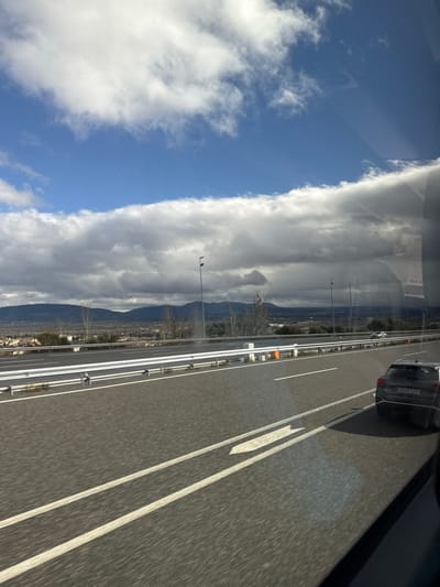 This image captures a highway scene from the perspective of a moving vehicle, looking out the passenger window. The road, featuring multiple lanes of asphalt, dominates the lower right portion of the frame. White lane markings, including solid lines, broken lines, and a large directional arrow, are clearly visible. A metallic guardrail separates the main lanes from the shoulder.

To the left of the road, the landscape unfolds towards distant mountains and hills, with sparse trees and some visible buildings or a small town nestled at their base. Utility poles line the roadside, with a small dark bird perched atop one.

The sky is a striking element, showcasing a clear blue area on the upper left, sharply contrasted by a large, elongated band of dense white and grey clouds stretching horizontally across the middle of the scene. This distinct cloud formation creates a dramatic visual division.

Other vehicles are present on the highway; a dark-colored car, possibly an SUV, is in the adjacent lane, displaying a European license plate that appears to read "8369 KJJ". Reflections of the interior of the vehicle from which the photo was taken, including some indistinct shapes and faint text, are visible superimposed on the sky in the upper frame.

The scene depicts a daytime setting with partly cloudy weather, suggesting a journey through a mountainous region. The highway infrastructure and landscape align with a location such as Guadarrama, Spain.