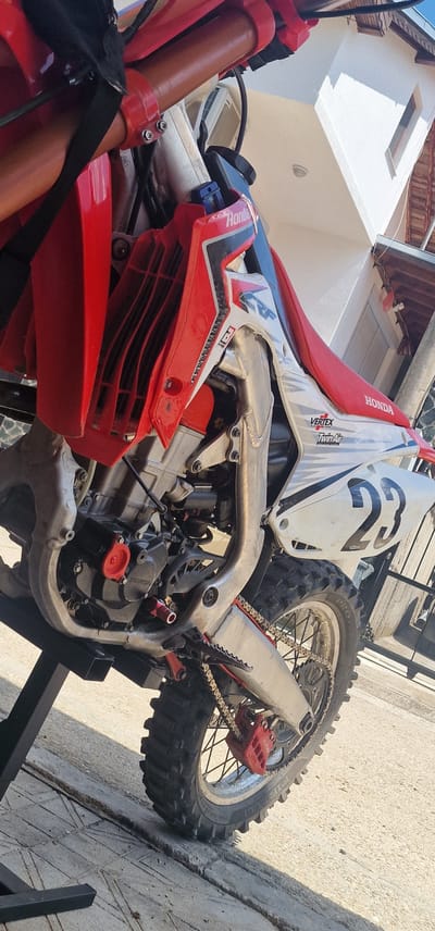 A red and white Honda CRF motocross motorcycle is prominently featured, resting on a black metal stand on a paved concrete surface. The bike exhibits red radiator shrouds with black vertical vents, a red textured seat, and white side panels marked with the black number '23'. Visible sponsor decals include 'Vertex,' 'TwinAir,' and a small 'FMF Racing' logo. The engine casing is black, bearing a 'Honda' emblem, with a distinct red cap on the side. The polished aluminum frame shows visible weld lines. The rear wheel is equipped with a knobby Pirelli tire, a silver rim, black spokes, and a red brake caliper. A silver drive chain is also visible. The upper left of the image shows red handlebars with black sleeves and a brown-orange front fork stanchion held by a red triple clamp. In the background, a multi-story white building with a small, dark-framed rectangular window and a dark-tiled roof is present, along with a partially visible outdoor air conditioning unit mounted on its facade. Bright sunlight illuminates the scene, casting pronounced shadows on the light grey concrete paving, which exhibits distinct seams and wear. The sky above is clear and blue. No individuals are depicted in the image. The scene is set in Kolarovo, Bulgaria.