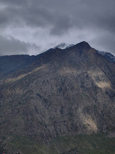 The image shows a mountainous landscape under a cloudy sky. The foreground is dominated by a rugged, dark-colored mountain slope with patches of green vegetation. A beam of light illuminates a section of the slope, highlighting its textured surface. In the background, another mountain range with snow-capped peaks is visible, partially obscured by clouds. The overall mood is somber and dramatic due to the heavy cloud cover and the imposing mountain terrain. The scene is likely located in the high-altitude region of Gondhla, India, given the characteristic geology and vegetation. There are no people or specific objects, except for what appears to be a small structure with a red roof visible at the very bottom left edge of the image.