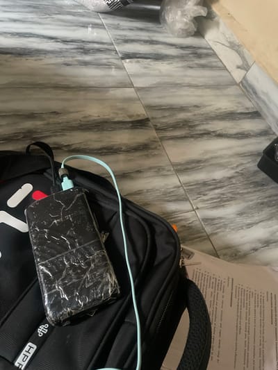 An overhead shot shows a black backpack lying on a marble-patterned floor. A power bank, wrapped in clear plastic, is on top of the backpack, connected by a light blue charging cable to a black cord. The backpack has white lettering on it, and a tag with black and white text reading "HPF" is visible near the bottom. A document with printed text is partially visible underneath the backpack. The floor tiles are a mixture of gray and white, with a vein-like pattern. The scene appears to be indoors, possibly in a home or office setting. No people or actions are depicted.