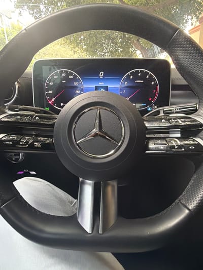 The image shows the interior of a Mercedes-Benz car, focusing on the steering wheel and dashboard. The steering wheel is black leather with silver accents and the Mercedes-Benz logo in the center. On the dashboard, there is a digital display showing a speedometer and a tachometer. The speedometer shows a reading of 0 mph, and the tachometer shows a reading of 0 x1000/min. There are also various buttons and controls on the steering wheel and dashboard. A light gray fabric is visible in the lower part of the image, likely part of the car's interior or a garment. The scene appears to be captured during the day, with natural light filtering through what appears to be trees outside the car. The location is identified as Pasadena, United States of America.