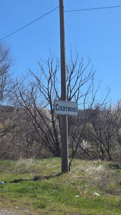 The image shows a sign for Slatino, Bulgaria. The sign is attached to a utility pole. The background consists of a clear blue sky, bare trees, and some green grass.