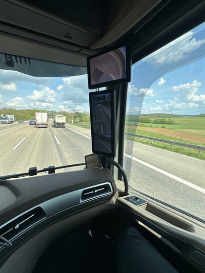 The image presents a first-person view from inside the cabin of a large vehicle, likely a truck, traveling on a multi-lane highway. The perspective is through the windscreen, extending upwards to a panoramic roof that reveals a bright blue sky dotted with fluffy white clouds.

Inside the cabin, elements of the dashboard, including air vents and a fragment of the steering wheel, are visible on the right. Two digital displays serve as virtual mirrors, mounted on the upper part of the windscreen frame. The left screen shows a curved road view, while the central screen displays the highway and vehicles behind the observer. A dark-clad leg of an occupant is partially visible on the far right. The interior design appears modern.

Outside the vehicle, a multi-lane highway stretches into the distance, marked with white lines. Other vehicles, including several trucks and cars, are visible on the road. A metal guardrail runs alongside the highway, separating it from expansive green fields and distant tree lines, indicative of a rural or semi-rural landscape. One truck in the distance has a partially legible name, possibly "Kreitzhel," on its side. The scene is brightly lit, suggesting clear, sunny daytime weather. The context places this scene in Reiskirchen, Germany.