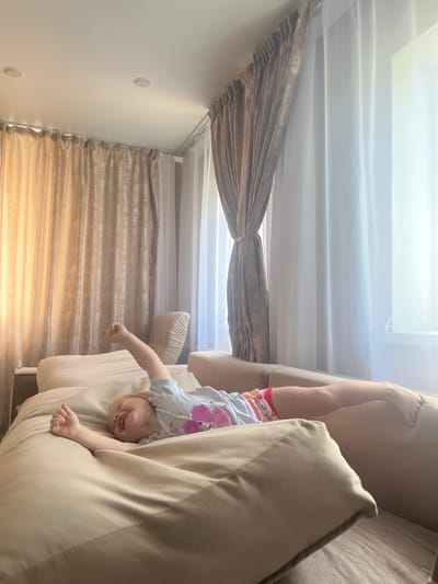 A young child with blonde hair is lying on a large, beige sofa, stretching their arms and legs. The child is wearing a light blue t-shirt with pink hearts and colorful shorts. The sofa is positioned in a room with large windows covered by beige and white curtains. The environment appears to be a residential interior, possibly a living room. The scene is brightly lit by natural light filtering through the curtains. There are no other people or animals visible in the image. The overall impression is one of a relaxed or playful moment. The location in Rakhinka, Russia, is not visually indicated in the image.