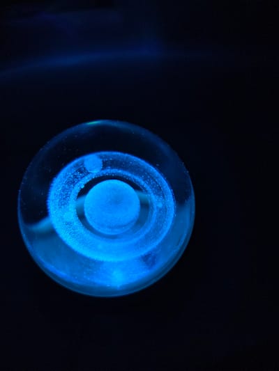 The image shows a glowing blue spherical object against a dark background. The object consists of concentric rings of light, with a central, brighter sphere. The outer ring is more diffuse. Small specks of light are scattered within the rings, giving them a granular appearance. A faint blue light source illuminates the left side of the frame. The image provides no information related to Cherkovitsa, Bulgaria, people, subjects, actions, interactions, or the environment.