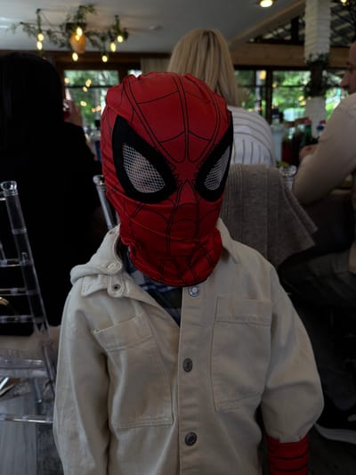 A person wearing a Spider-Man mask is sitting indoors, visible from the shoulders up. They are wearing a light beige or white jacket with visible pockets and button details. The jacket is worn over a patterned shirt. The environment includes other people partially visible, along with decorations and what appear to be windows in the background, but the image does not show any information or geographical features that allows to identify the location as Lermontovo, Armenia. There is nothing in the image to indicate Lermontovo, Armenia or any other city.