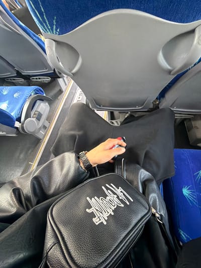 Inside a vehicle, possibly a bus, a person's hand is visible holding a small object. The person is wearing a watch and a black leather jacket. A black bag with white writing is positioned next to the jacket. A black bag rests on the floor. Seating with blue fabric is visible. The location is not discernible from the image.