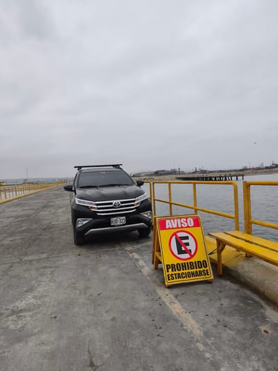 An outdoor wide shot captures a black Toyota Rush SUV parked on a concrete pier or dock under an overcast sky in Chimbote, Peru. The vehicle, equipped with roof rails, faces the viewer slightly angled to the right. Its front license plate, "PERU H3D-321", is clearly visible, along with a "TOYOTA Rush" and "Autonort" label.
To the right of the car, a prominent yellow A-frame sign with red lettering stands on the pier. The sign reads "AVISO" (Notice/Warning) at the top, followed by a universal "No Parking" symbol (a red circle with a diagonal line through a black 'E'), and "PROHIBIDO ESTACIONARSE" (No Parking) at the bottom. Despite the warning, the car is parked directly in front of the sign.
Beyond the sign, a bright yellow metal railing runs along the edge of the pier, separating it from the calm, dark grey water. A matching yellow wooden bench is positioned against the railing. In the distance, across the water, the coastline of Chimbote is visible, featuring various industrial structures, buildings, and possibly ships or cranes, indicating a port environment. The sky is uniformly cloudy, suggesting an overcast day with diffused light. No people are visible in the image.