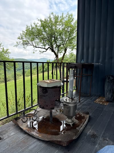 The image depicts an outdoor setting, likely a balcony or porch, overlooking a lush green landscape with rolling hills and trees under a cloudy sky. In the foreground, there are several objects associated with outdoor cooking and hospitality. A large, rustic metal brazier sits on a weathered metal tray that has collected some water. On top of the brazier, there is a light-colored box containing what appear to be wooden skewers or utensils. To the right of the brazier is a traditional metal samovar, a device for heating and boiling water for tea, also situated on the metal tray. Behind the samovar and brazier, a rusty metal grill stand holds a grill grate and a single skewer. A dark-colored, ribbed wall, likely part of a building, forms the backdrop on the right side of the image. A black plastic bag is visible near the wall. The ground surface appears to be made of dark, slatted material. The scene is set in the city of Sis, Azerbaijan. No people are visible in the image.