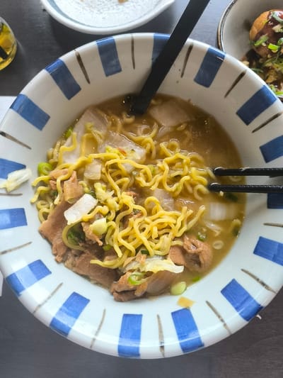 The image shows a bowl of ramen with broth, noodles, meat, and vegetables. Chopsticks and a spoon rest on the rim of the bowl. The bowl is white with a blue and white geometric pattern. The ramen is likely served in a restaurant.
While no other information in the picture points to the city of Leidschendam, Netherlands, the location is added to the prompt.