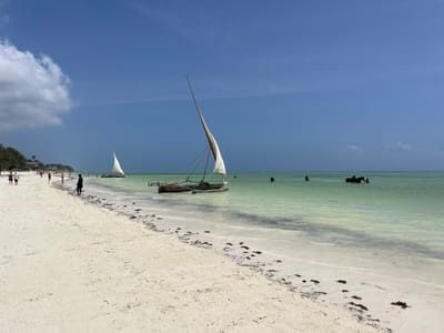 This image depicts a beach scene in Paje, Tanzania, under a clear blue sky with scattered white cumulus clouds primarily on the left. The foreground comprises a wide expanse of light-colored sand, appearing white or very light tan, marked with patches of dark organic material, likely seaweed, along the waterline and extending onto the dry beach. The ocean water, extending from the shore to the horizon, is a light greenish-blue, indicating shallow depths near the beach.

Approximately 15-20 individuals are present. Multiple figures are walking along the distant left shoreline. A single person in dark clothing stands at the water's edge to the left of the primary boat. Several individuals are partially submerged in the water to the right, with at least two positioned near a dark, partially submerged object or vessel.

Two traditional wooden outrigger canoes, each equipped with a triangular (lateen) sail, are prominent in the water. One canoe, larger and more defined, is positioned slightly right of the frame's center in the shallow water, its sail fully deployed. A second, smaller canoe with a deployed sail is situated further out and to the left of the first. Distant green vegetation lines the beach on the far left. The scene is illuminated by strong, direct sunlight.