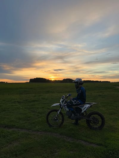 A dirt biker and his motorcycle are positioned on a grassy field. The sunset is visible in the distance, divided by a treeline. The sky displays varying shades of gray, blue, and orange. The biker is wearing a helmet, goggles, and riding gear. The location is near Новочувашский, Russia, indicated by coordinates.
