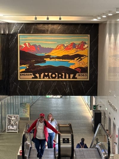The image is a composite of two photographs. The top image shows a large, framed poster of a landscape with mountains and a lake, set against a dark, marbled wall. Text below the image reads "ENGADIN ST. MORITZ 1856". The bottom image shows an interior scene with people on escalators. In the foreground, a man in a red jacket and baseball cap, and a woman in a blue shirt and jeans are descending on an escalator. Another person, wearing a backpack, is ascending on a separate escalator in the background. Several pieces of artwork are displayed on the walls. The location is identified as the city of Sesto San Giovanni, Italy.