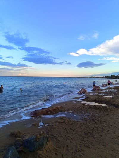 A wide-angle, low-angle shot captures a serene beach scene on a warm day at Lake Kulunda, located in Yarovoye, Russia. The image is divided into three horizontal bands: the sky, the sea, and the shore. The sky, occupying the top third of the frame, is a brilliant blue with scattered white and gray clouds. The sea, filling the middle section, is a deep blue with gentle waves lapping the shore. Several people are visible in the water, some swimming and others standing. The shore, the bottom third, is a mix of dark sand and scattered rocks, with some areas of shallow water reflecting the sky. The lighting suggests it is late afternoon or early evening, with the sun casting a soft glow on the scene. The overall impression is one of peaceful recreation and natural beauty.