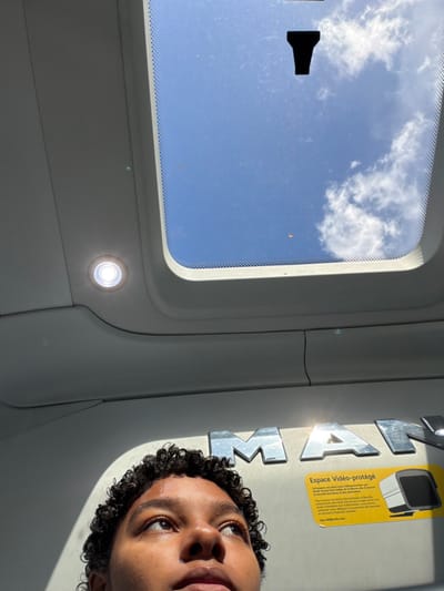 The image provides an upward perspective from within the passenger compartment of a Keolis public transport vehicle, likely a MAN model bus or coach, operating in the Île-de-France region of France, specifically implied to be in or around Noisiel.
The upper section of the frame shows a rectangular sunroof revealing a bright blue sky with scattered white cumulus clouds. A small, dark, inverted T-shaped object is adhered to the sunroof glass, which also displays minor surface debris or scratches and a small, blurred orange element. Below the sunroof, the vehicle's light-colored ceiling is visible, featuring an illuminated circular LED light fixture on the left. Prominently displayed on the ceiling are large, metallic, reflective letters spelling "MAN." To the right of these letters, a yellow rectangular sticker is affixed. This sticker, with text in French, designates the area as "Espace Vidéo-protégé" (Video-protected area) and includes contact information, specifically "dpo.idf@keolis.com" and the company name "Keolis," confirming its operational context within Île-de-France. The lower portion of the image shows the top of a person's head and face, with dark, curly hair and brown skin. The individual is looking upwards towards the sunroof, with direct sunlight casting highlights across their forehead and nose.