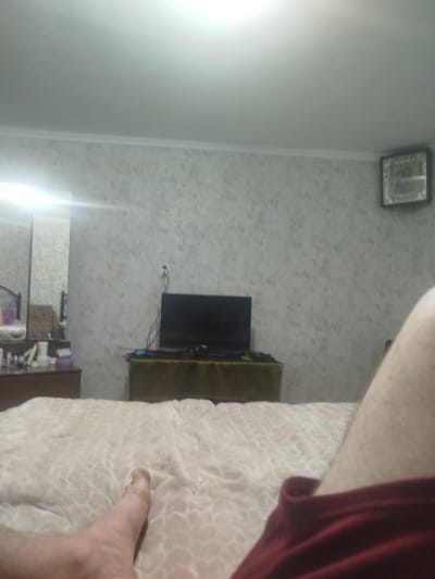 The image depicts an indoor scene, likely a bedroom. A large, flat-screen television is positioned on a dark-colored cabinet against a textured, light-colored wall. To the left, a dresser with various items on top, including bottles and containers, is visible next to a mirror. The foreground is dominated by a bed with a patterned, light-colored comforter. A person's legs and foot, clad in dark red shorts, are prominently featured in the lower right and bottom left of the frame respectively, suggesting the photo was taken from a reclining position on the bed. The lighting appears to be overhead, casting a bright glow on the ceiling. There are no discernible elements in the image that would indicate the specific location of Tomai, Moldova.